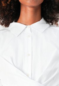 Camicia bianca con chiusura a bottoni, caratterizzata da una texture liscia e tessuto morbido, dettagliata con spalle ripiegate e un design pulito.