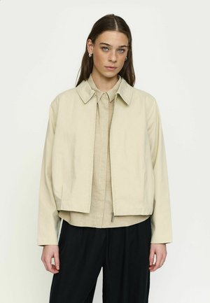 Noor naine, kellel on sirged pruunid juuksed, kandes beežist tõmblukuga jopet beeži särgi ja mustade pükstega, seismas ühevärvilise tausta ees.