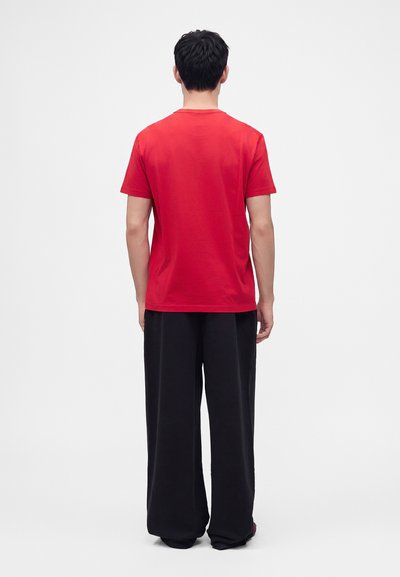 Homme debout, dos tourné, portant une chemise rouge unie à manches courtes et un pantalon noir ample, sur un fond clair uni.