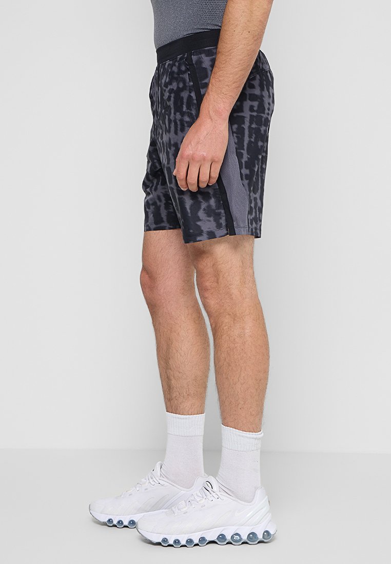 Homme portant un short de sport à motif gris, un t-shirt gris, des chaussettes blanches et des baskets blanches, se tenant de côté.
