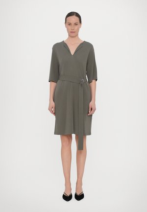 Woman standing wearing a knee-length olive green dress with elbow-length sleeves and a matching belt, paired with black pointed-toe heels.