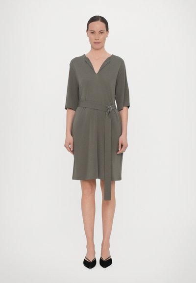 Vestido verde oliva con escote en V y mangas tres cuartos, ceñido en la cintura con un cinturón de tela que tiene una hebilla circular.