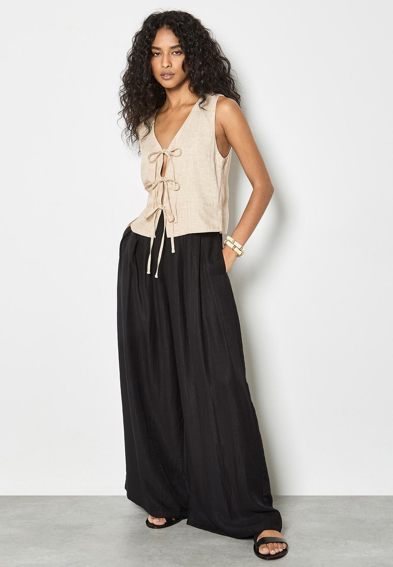 Mujer con cabello rizado, vestida con una blusa sin mangas beige con lazos al frente, pantalones anchos negros, sandalias y pulseras, sobre un fondo blanco.