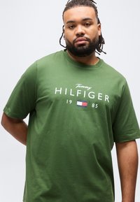 Άνδρας με πλεγμένα μαλλιά και γένια, φορώντας πράσινο T-shirt Tommy Hilfiger με λογότυπο και σημαία, σε λευκό φόντο.