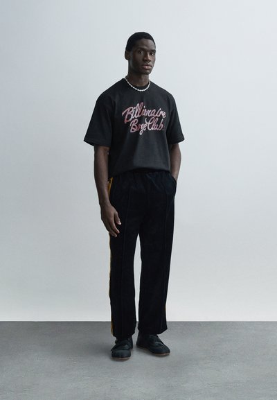 Μαύρο βαμβακερό μπλουζάκι με πολύχρωμο λογότυπο "Billionaire Boys Club", σε συνδυασμό με μαύρο βελούδινο παντελόνι με κίτρινες πλαϊνές ρίγες. Μαύρα αθλητικά παπούτσια.