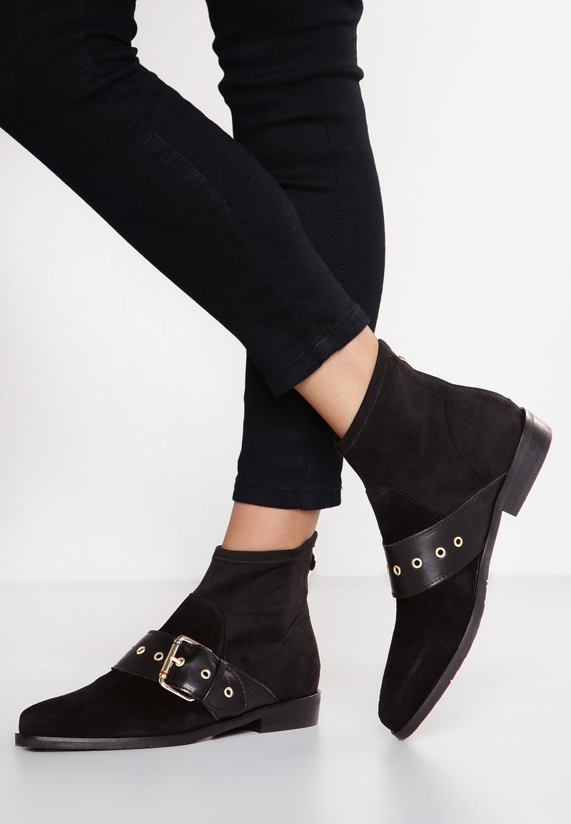 Botines de ante negros con una amplia correa de cuero que presenta ojales dorados y una hebilla decorativa. Tacón bajo y silueta elegante.
