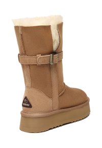 Brown suede ankle boot with a shearling lining, adjustable strap, and rubber sole. Features a triangular logo on the heel.