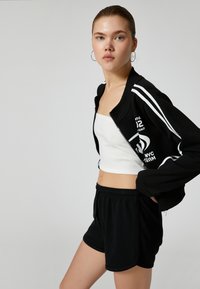 Sort zip-jakke med hvide striber og grafik, sammen med en hvid ribbet crop top og sorte shorts med snor. Glat tekstur.