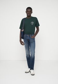 Zelené bavlněné tričko s grafickým logem, modré džíny a šedé tenisky. Model stojí s modrou taškou přehozenou přes jedno rameno.