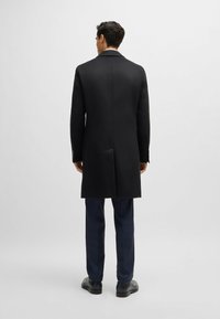 Cappotto lungo in lana nera con un design strutturato. Presenta rever a punta, uno spacco singolo sul retro e dettagli delle maniche su misura.