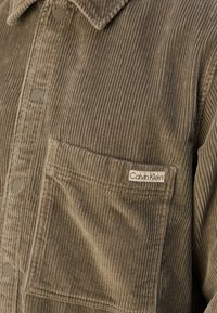 Corduroy jacket in olive green with a textured surface, featuring a chest pocket and a Calvin Klein label. Snap buttons along the front.