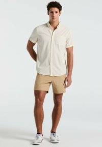 Licht beige kortefris overhemd met knoopsluiting, gemaakt van textuurstof en een enkele borstzak, gecombineerd met khaki shorts en witte sneakers.