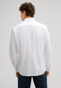 Camicia bianca a maniche lunghe con orlo dritto, polsini a bottone e una finitura leggermente testurizzata. Collo e cuciture in contrasto.
