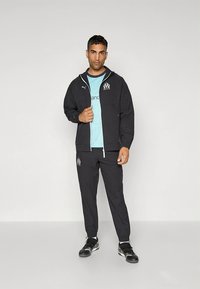 Must musta spordijakk kapuutsi ja lukuga, millel on logo; helesinine t-särk all; sobivad mustad püksid; mustad kingad valgete aksentidega.