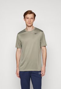 Tricou Nike de culoare verde măsliniu, cu guler rotund, mâneci scurte și un logo negru pe pieptul stâng. Țesătura pare netedă și ușoară.