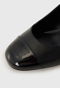 Scarpa nera con tacco alto, punta in vernice lucida, corpo in pelle opaca e dettagli cuciti lungo i bordi.