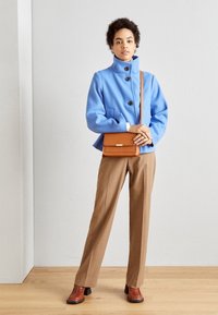 Jasnoniebieska kurtka z wysokim kołnierzem i czarnymi guzikami; brązowe, dopasowane spodnie; brązowa torebka; bordowe botki na obcasie; neutralne tło.