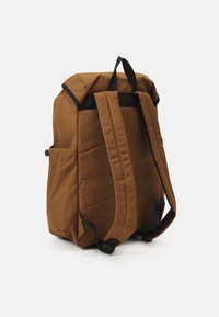 Mochila de tela marrón con textura acolchada, correas negras, bolsillo exterior y un diseño minimalista. La forma es rectangular con esquinas redondeadas.
