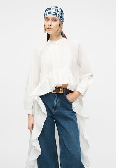 Γυναίκα που φοράει λευκό μπλουζάκι με μακριά μανίκια και βολάν, φαρδιά τζιν μπλε παντελόνια, καφέ ζώνη και μπλε μαντήλι με σχέδιο στο κεφάλι, ποζάροντας με το ένα χέρι στην τσέπη.