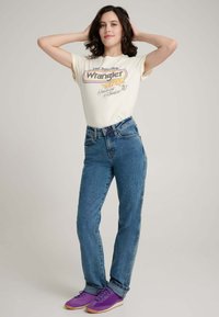 Cremfarbenes T-Shirt mit retro "Wrangler"-Grafik, kombiniert mit blauen, hochtaillierten Jeans und lila Turnschuhen. Schlichter Hintergrund.