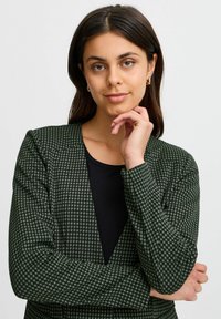 Zaļa houndstooth raksta žakete ar nedaudz strukturētām plecu daļām, garām piedurknēm un dziļu V-veida kaklu virs melna krekla.