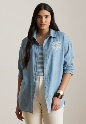 Femeie cu păr lung și închis la culoare poartă o cămașă albastru deschis cu nasturi peste un top în dungi, pantaloni albi și brățări aurii și de tip scoică țestoasă, stând în fața unui fundal bej.