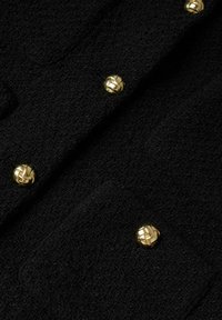Sort tekstureret stof med skinnende guld runde knapper med et vævet mønster, der er arrangeret lodret langs beklædningen.