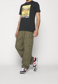 Svart grafisk T-shirt med ett gult bergtryck, kombinerad med olivgröna cargobyxor med sidofickor och svart-röda sneakers.