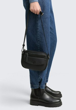 Borsa a tracolla nera in pelle con finitura testurizzata, compartimenti con zip e tracolla regolabile, tenuta da una persona che indossa jeans blu e stivali neri.