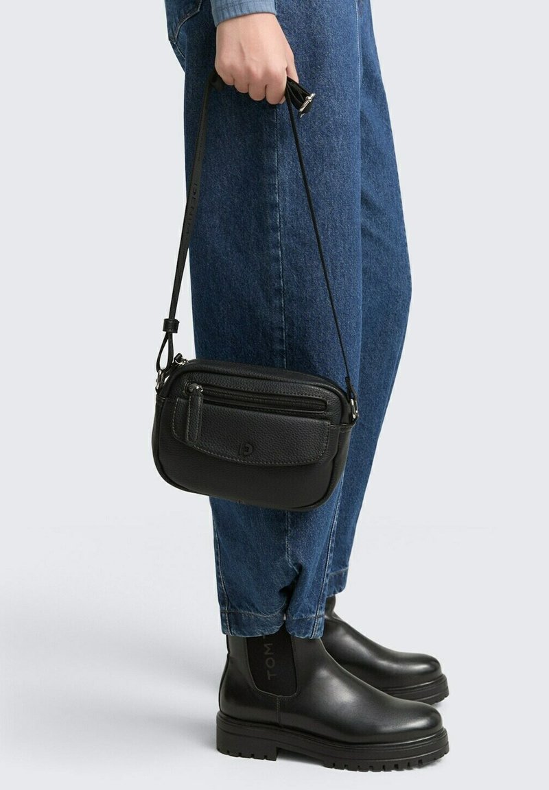 Schwarze Leder-Crossbody-Tasche mit strukturiertem Finish, Reißverschlussfächern und einem verstellbaren Trageriemen, gehalten von einer Person, die blaue Jeans und schwarze Stiefel trägt.