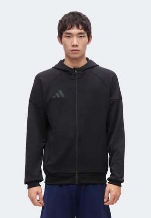 Jeune homme portant une veste à capuche noire zippée avec un logo subtil, debout face à l'avant sur un fond clair uni.