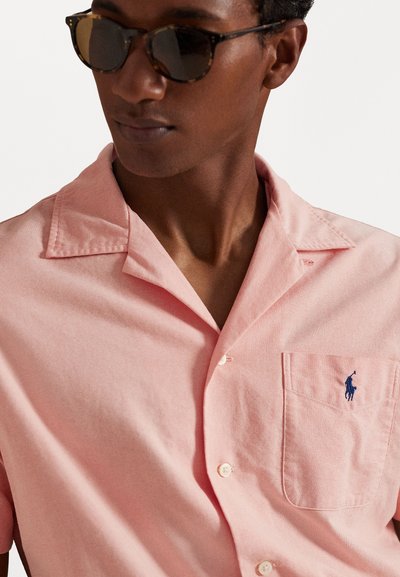 Φανελάκι κοντού μανικιού, σε ανοιχτό ροζ χρώμα, από βαμβάκι, με τσέπη στο στήθος και ένα μικρό μπλε λογότυπο στην τσέπη. Κλασικός σχεδιασμός για λαιμό.