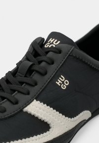 Sapatilha desportiva preta com parte superior em couro texturizado, detalhes em camurça bege, atacadores planos pretos e o logótipo "HUGO" em dourado na língua.
