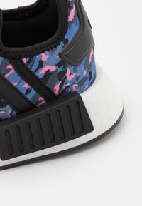 Sportskor med ett svart och mångfärgat kamouflagemönster, tålig tygöverdel, tjock vit mellansula med texturerade detaljer och svart yttersula.