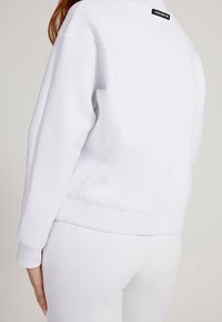 Biała bluza o luźnym kroju, z okrągłym dekoltem i ściągaczami. Posiada małą czarną metkę z tyłu dekoltu. Gładka struktura.