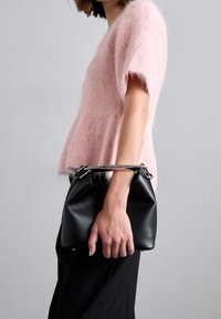 Černá kožená kabelka s kulatou základnou, stříbrným kováním a strukturovaným designem, držena v ruce. Nosená s jemně růžovým chlupatým topem.