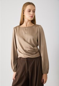 Beige wikkeltop met lange mouwen en een ronde halslijn, voorzien van een zachte textuur en subtiele plooien bij de taille. Gecombineerd met bruine broek.