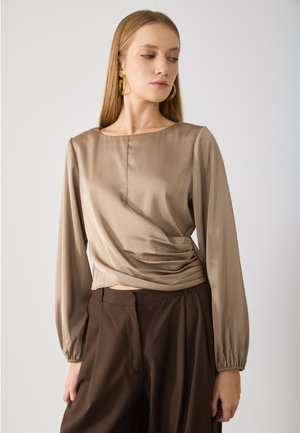 Beige Wickeloberteil mit langen Ärmeln und rundem Halsausschnitt, das eine glatte Textur und dezente Raffung an der Taille aufweist. Kombiniert mit braunen Hosen.