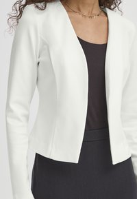 Veste blanche ajustée en tissu lisse, dotée de manches longues, d'un devant ouvert, et d'une silhouette élégante, associée à un haut foncé.