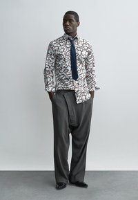 Man staat met zijn handen in zijn zakken, gekleed in een lange mouw overhemd met patroon, een marineblauwe stropdas, losse grijze broek en zwarte schoenen tegen een effen achtergrond.