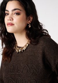 Bruine gebreide trui met textuurrijke letters "MILAN" op de voorkant, aangevuld met een gouden statementketting met verschillende hanger-vormen.