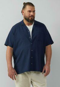 Granatowa koszula z krótkim rękawem w delikatny wzór w kratkę, z kieszenią na lewej klatce piersiowej i guzikami z przodu. Nosi się ją na białym T-shircie.
