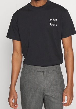 Mann, der ein schwarzes T-Shirt mit kurzen Ärmeln und weißem Aufdruck „AVANT - APRÈS“ auf der linken Brust trägt, kombiniert mit grau strukturierten Hosen.