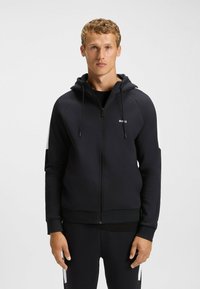 Black zip-up hoodie with a fitted design, soft fabric, and a white logo on the chest. Features adjustable drawstrings and contrasting shoulder accents.