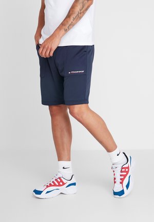 Mężczyzna noszący granatowe szorty, białe skarpetki Nike oraz białe buty sportowe z niebieskimi i czerwonymi akcentami, stojący z rękami w kieszeniach.