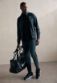 Træningstøjet inkluderer en mørkegrøn zip-up jakke, en sort t-shirt, mørkegrønne joggers og sorte sneakers. En stor grå Nike weekendtaske.