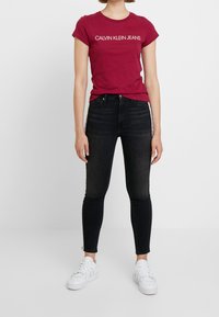 Camiseta roja de cuello redondo con "CALVIN KLEIN JEANS" impreso en blanco, combinada con jeans skinny negros y zapatillas blancas. Diseño minimalista.