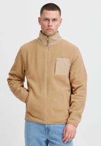 Tan fleece jacket with a high collar, zip closure, and a front pocket. Soft texture, relaxed fit, featuring side pockets.