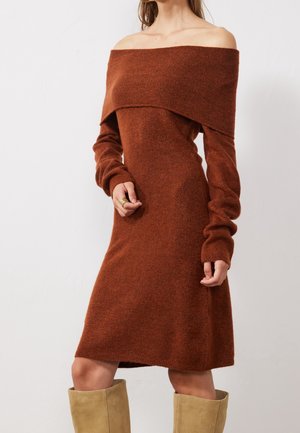 Vrouw draagt roestkleurige off-shoulder gebreide jurk met lange mouwen en beige kniehoge laarzen, staand voor een effen witte achtergrond.