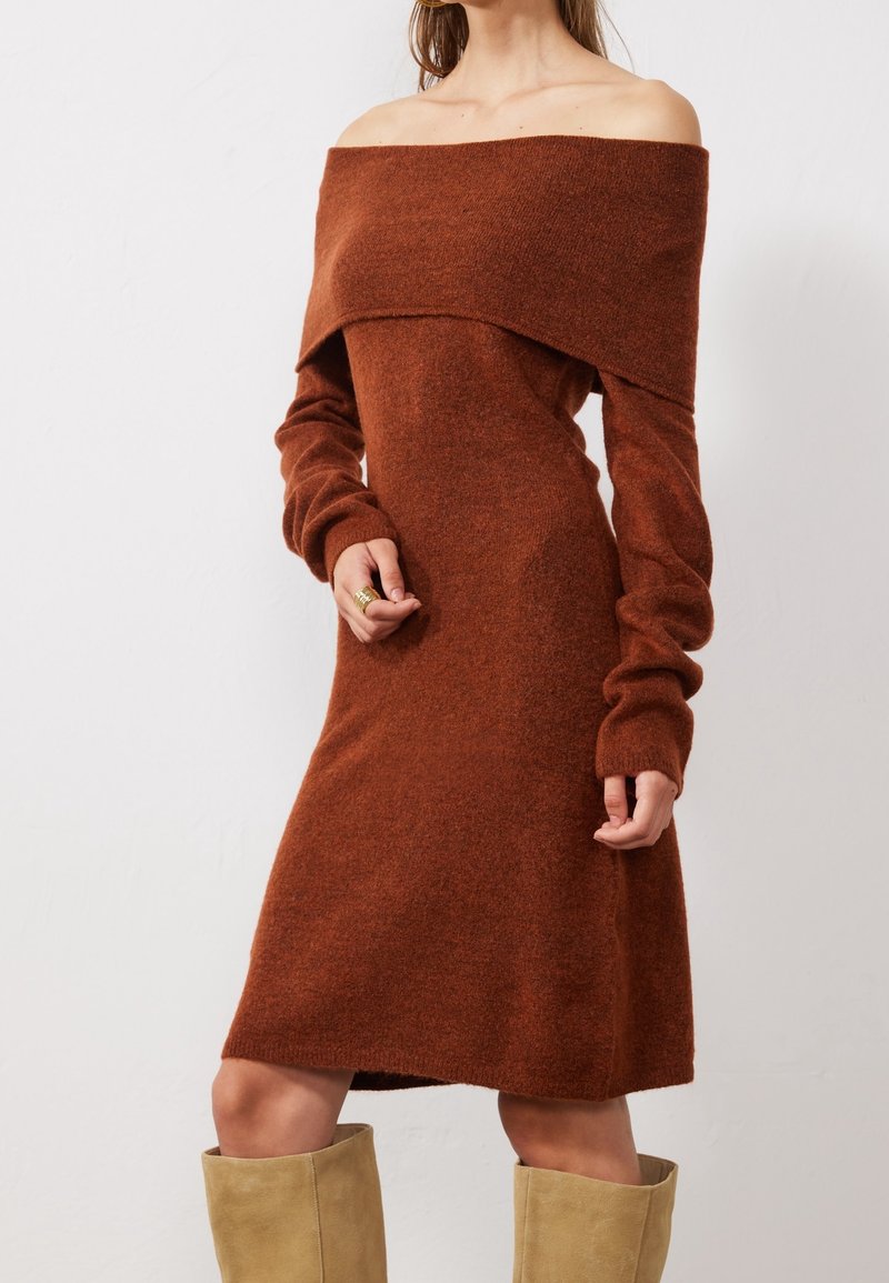 Femme portant une robe en maille couleur rouille, épaules dénudées, manches longues, et des bottes beige montant jusqu'aux genoux, debout devant un fond blanc uni.